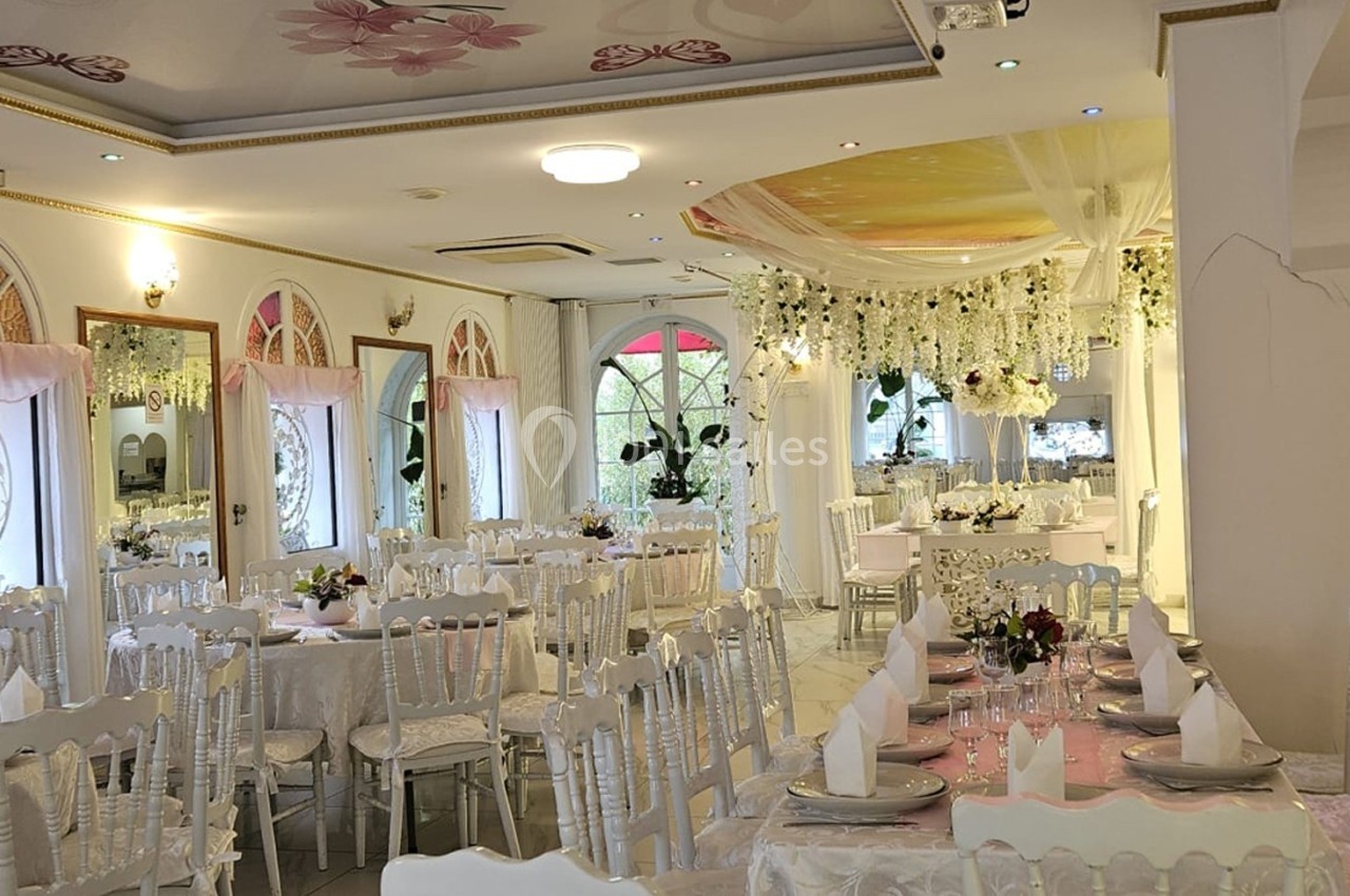 Salle de réception lumineuse décorée de fleurs blanches, tables dressées avec nappes blanches et chaises élégantes.