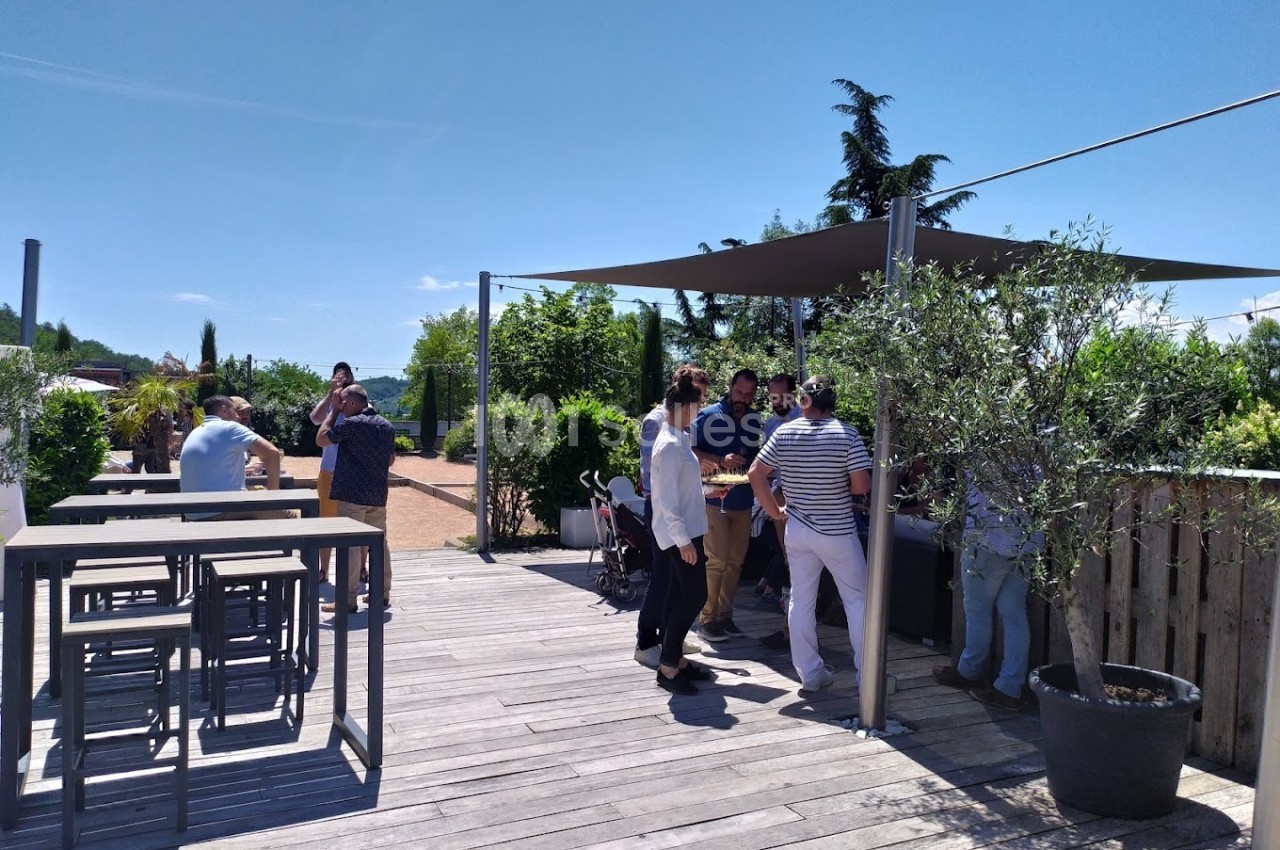 Personnes discutant sur une terrasse en bois ombragée, entourée de végétation, par une journée ensoleillée.