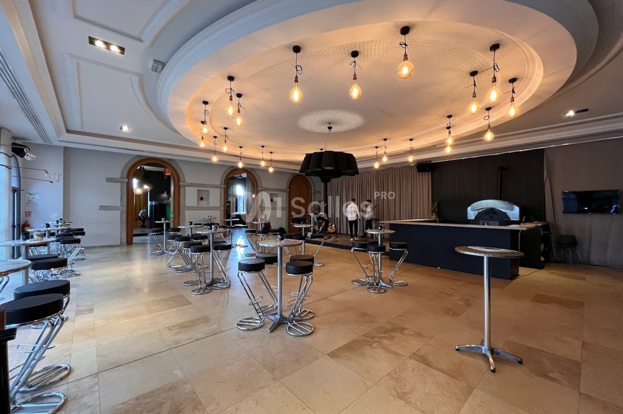Salle lumineuse avec un plafond orné de luminaires, des tables hautes et un comptoir moderne au fond.