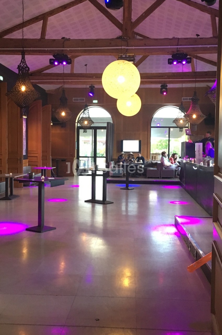 Salle avec un bar, tables hautes et éclairage violet, grandes fenêtres ouvertes sur une terrasse.