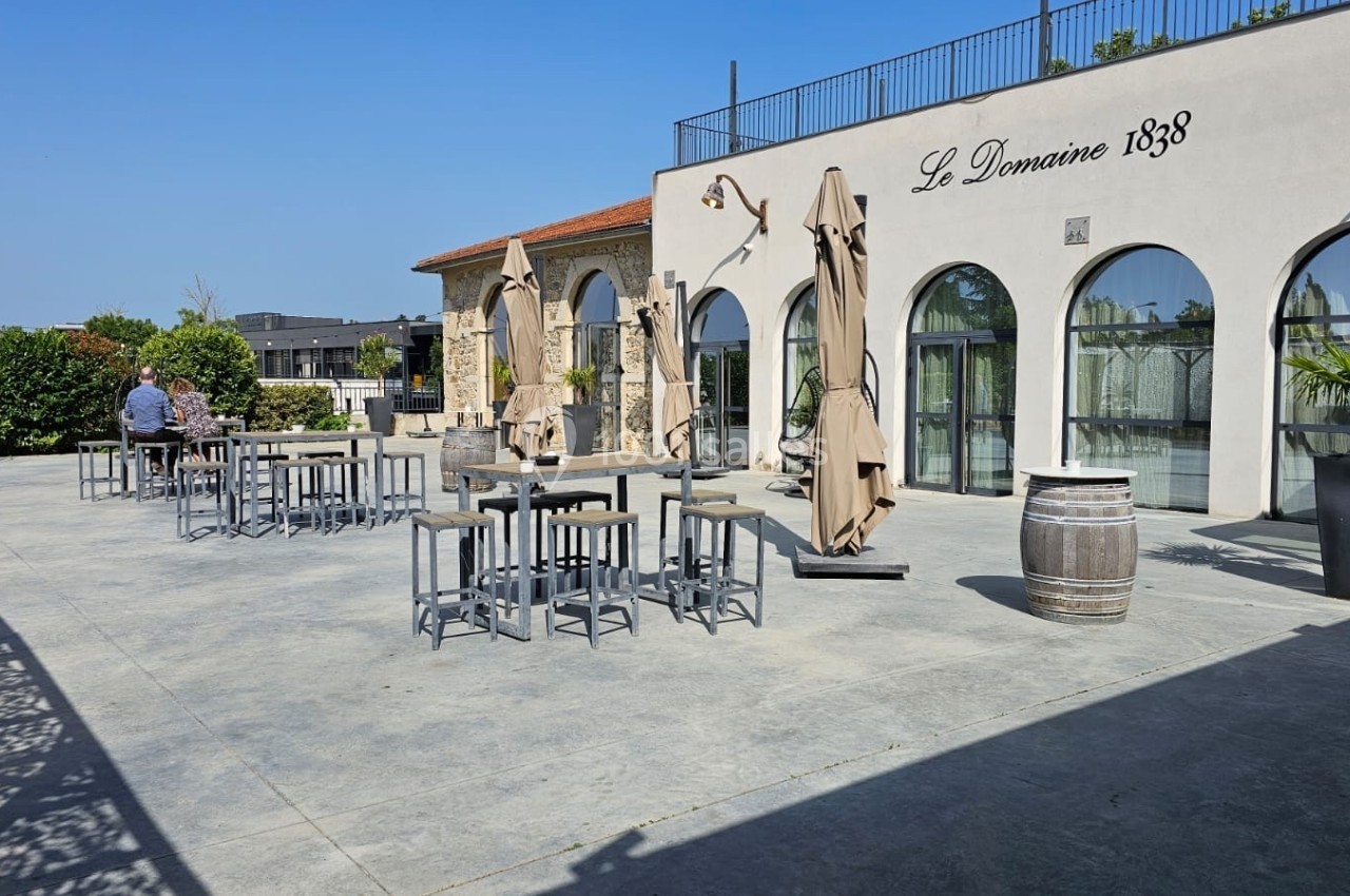 Terrasse ensoleillée avec tables hautes, parasols fermés et façade d'un bâtiment marqué ’Le Domaine 1838’.