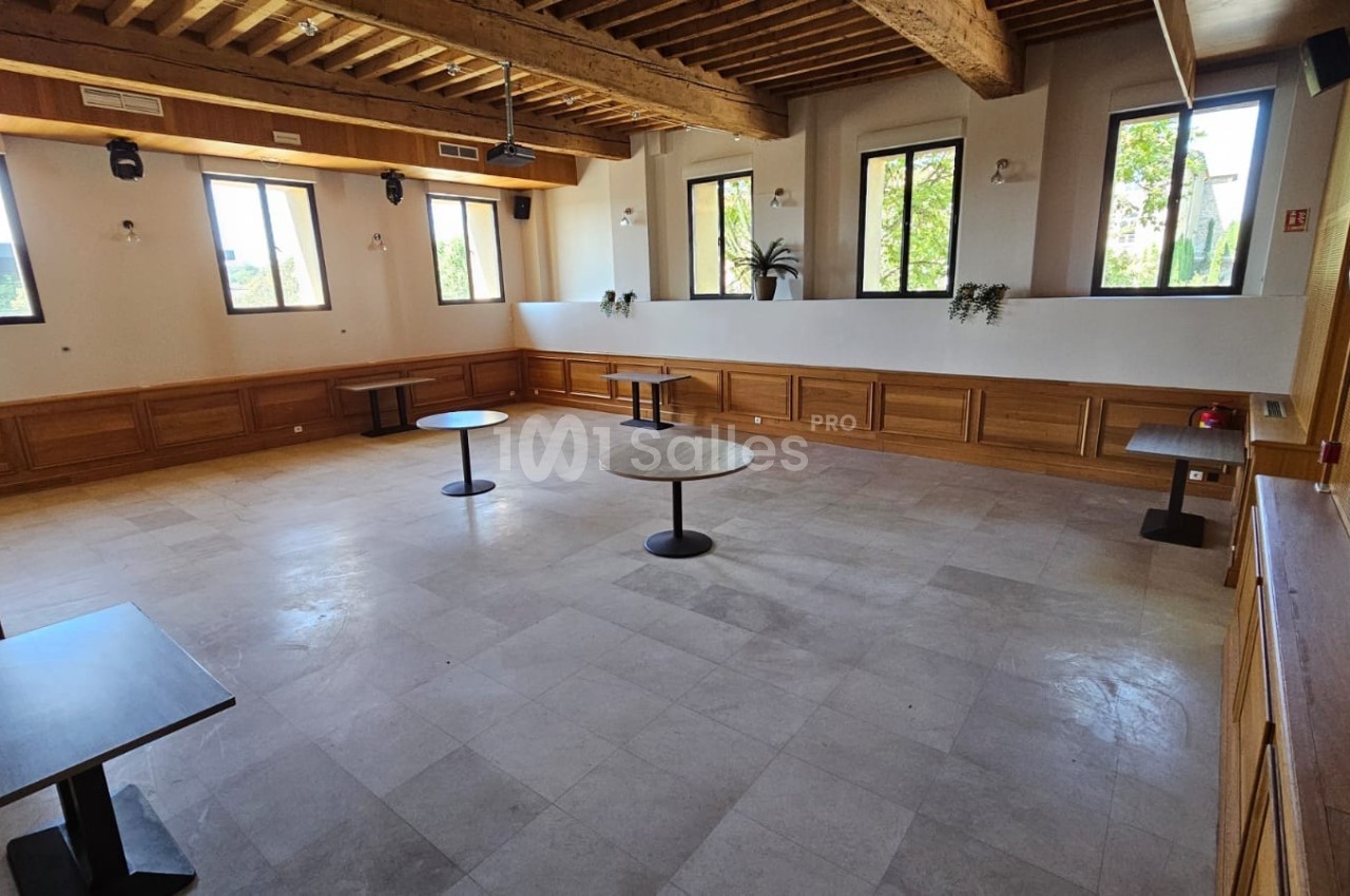 Salle lumineuse avec parquet clair, poutres apparentes, tables rondes espacées et grandes fenêtres donnant sur l'extérieur.