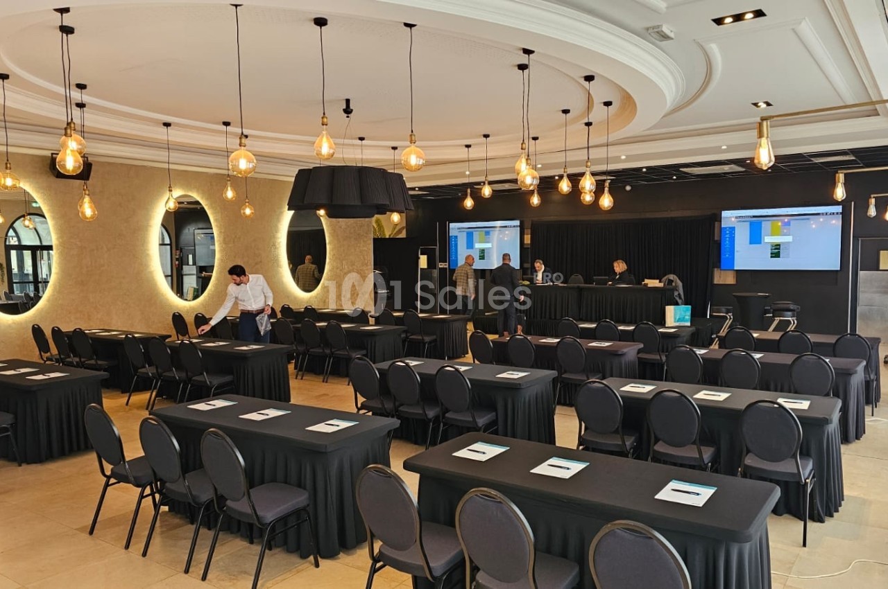 Salle de conférence avec des chaises alignées, des tables noires, des écrans en arrière-plan et des lumières suspendues.