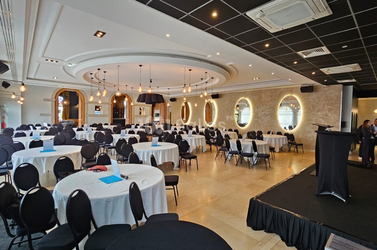 Salle de réception élégante avec tables rondes dressées, chaises noires et une estrade équipée d'un pupitre.