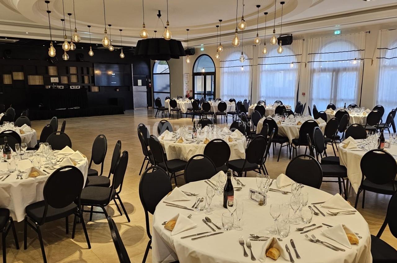 Salle de réception avec tables rondes dressées, nappes blanches, chaises noires et éclairage suspendu.