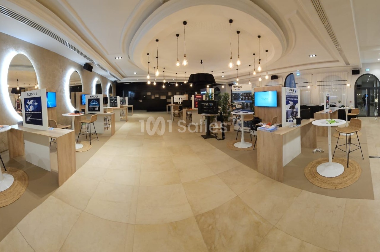 Salle d'exposition lumineuse avec stands, écrans et tables hautes dans un espace moderne et épuré.