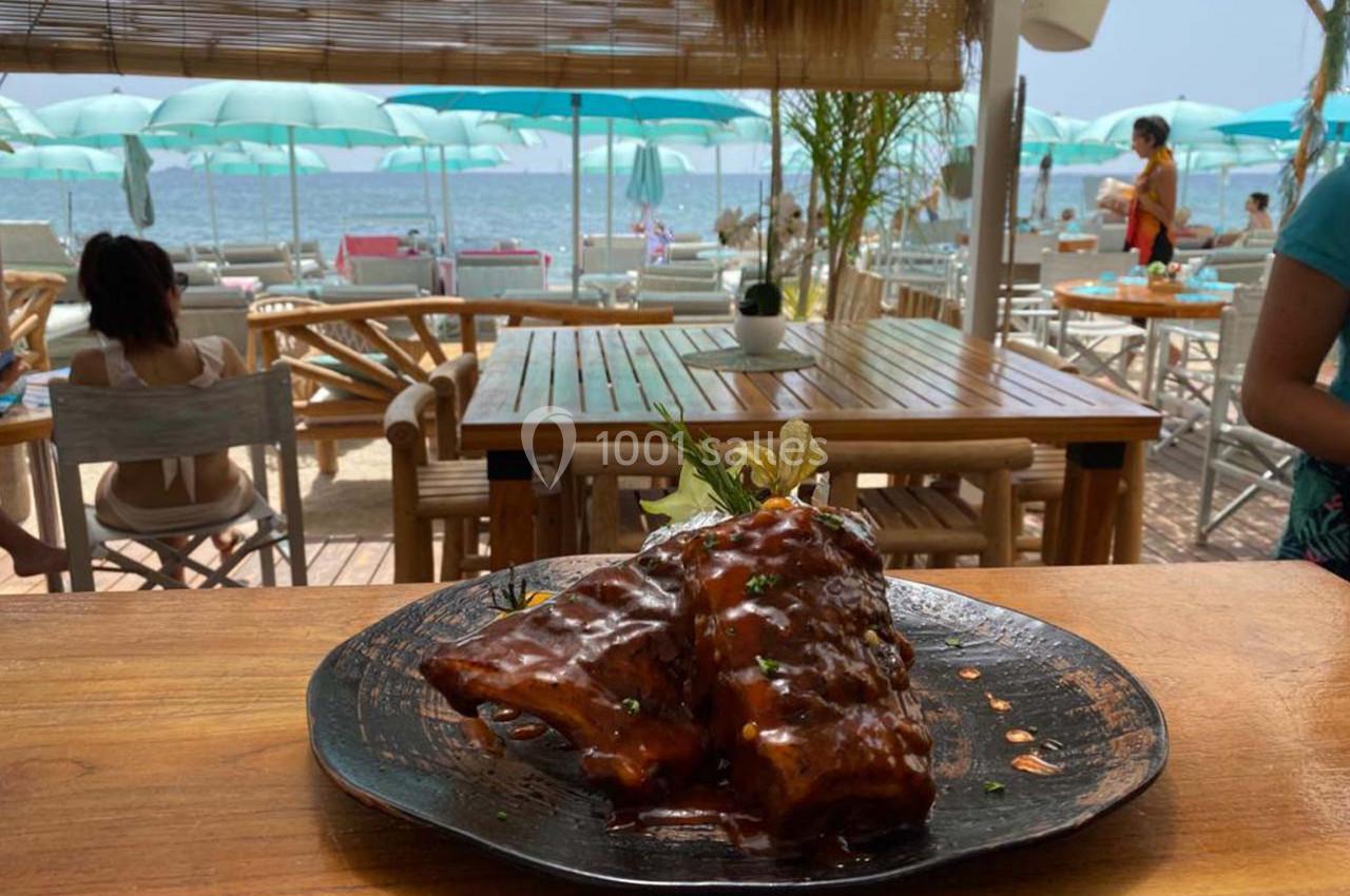 Assiette de côtes de porc grillées avec sauce, servie sur une terrasse en bois face à une plage avec parasols.