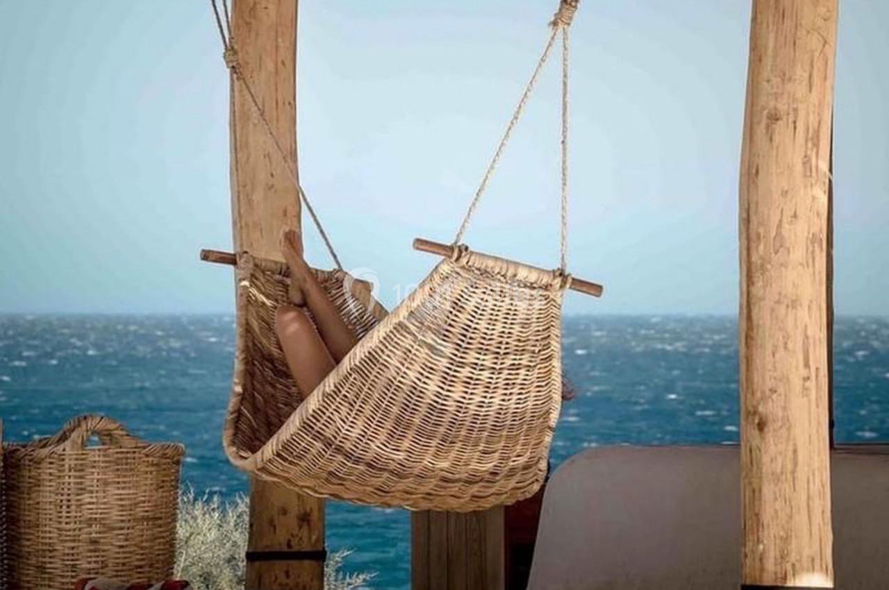 Hamac en osier suspendu sous une pergola en bois, avec vue sur la mer et un panier en osier à proximité.