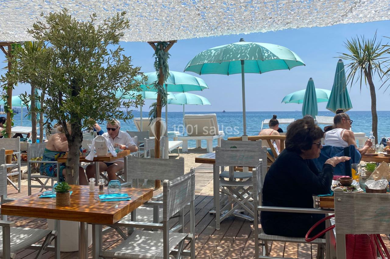 Terrasse d'un restaurant en bord de mer avec des parasols turquoise, des tables en bois et des clients attablés.