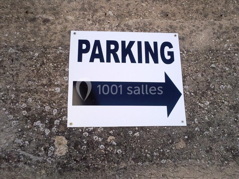 Panneau blanc avec le mot ’PARKING’ en noir et une flèche noire pointant vers la droite, fixé sur un sol en béton.