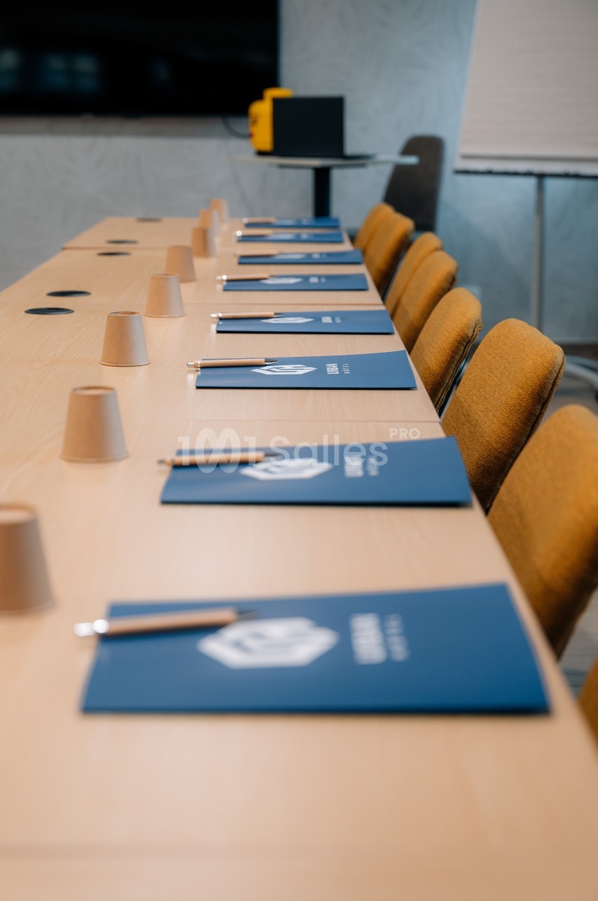Salle de réunion avec une table équipée de carnets bleus, stylos et gobelets en carton alignés devant des chaises jaunes.