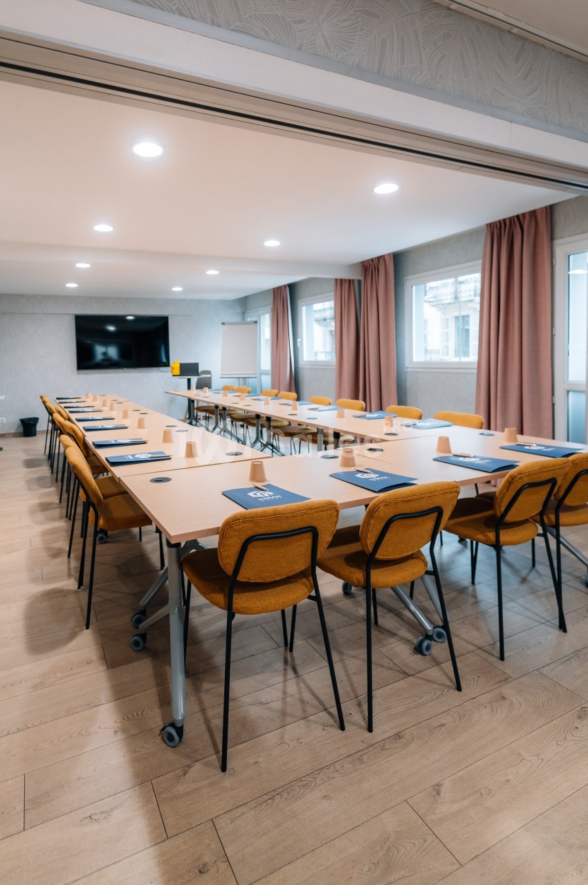 Salle de réunion lumineuse avec de longues tables, des chaises jaunes, un écran mural et des rideaux roses.