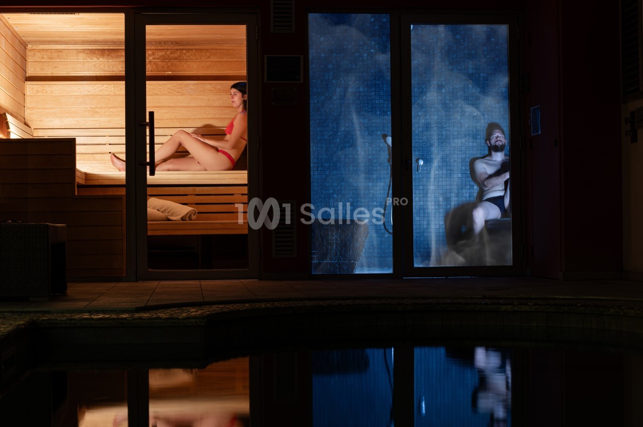 Une femme dans un sauna en bois et un homme dans un hammam vitré avec de la vapeur, vue depuis l'extérieur.