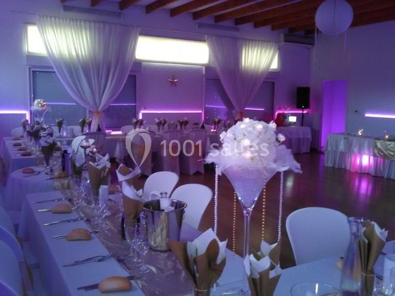 Salle de réception décorée avec des tables dressées, nappes blanches, centres de table fleuris et éclairage violet.
