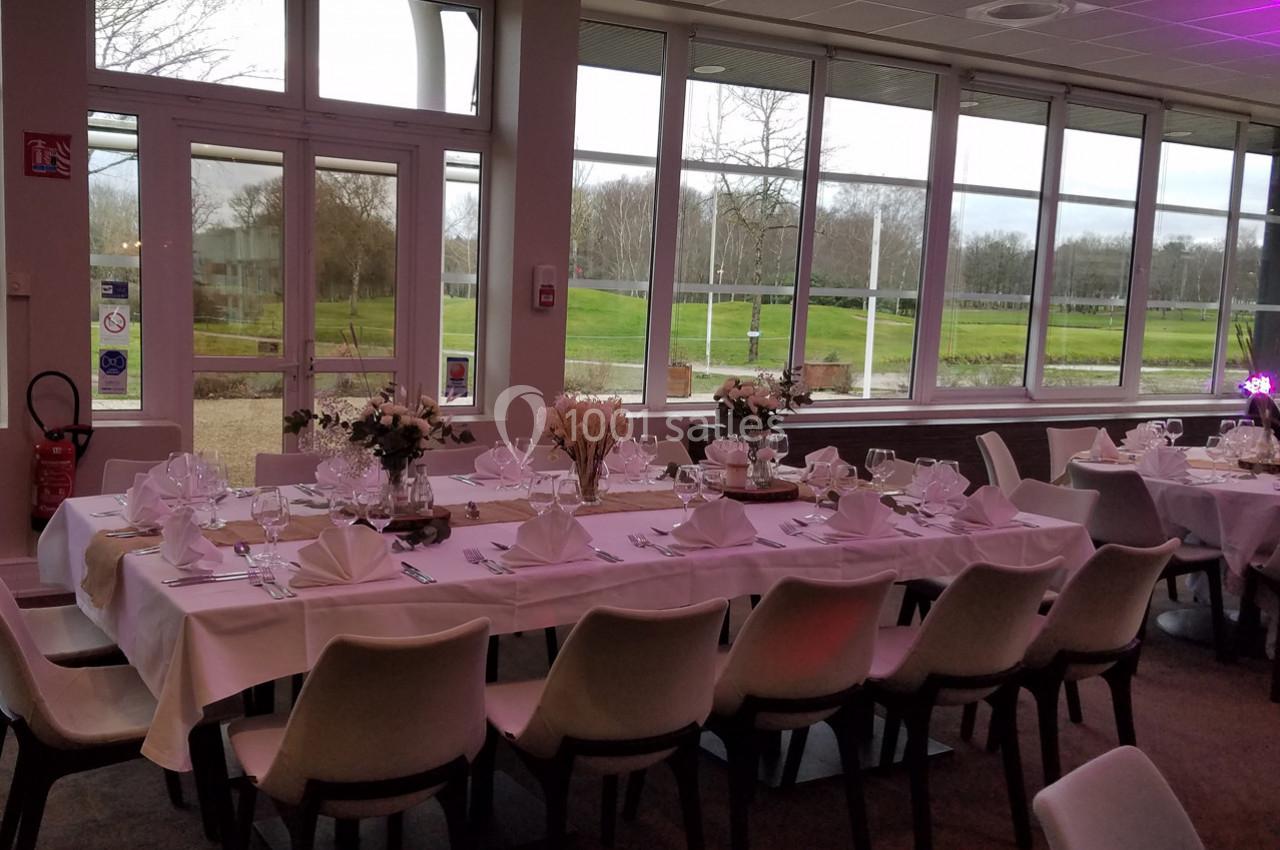 Salle de réception avec tables dressées, nappes blanches, décorations florales et vue sur un extérieur verdoyant.