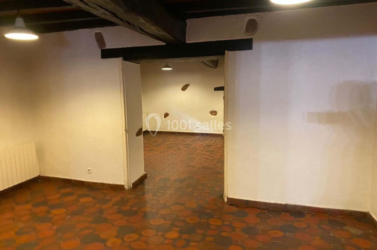 Salle vide avec carrelage en terre cuite, murs blancs, poutres apparentes au plafond et éclairage suspendu.