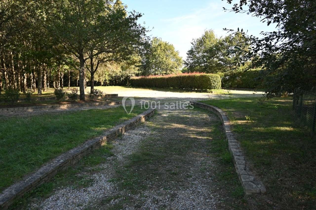 Allée en gravier bordée de pelouses et d'arbres, menant à une haie et une clairière ensoleillée.