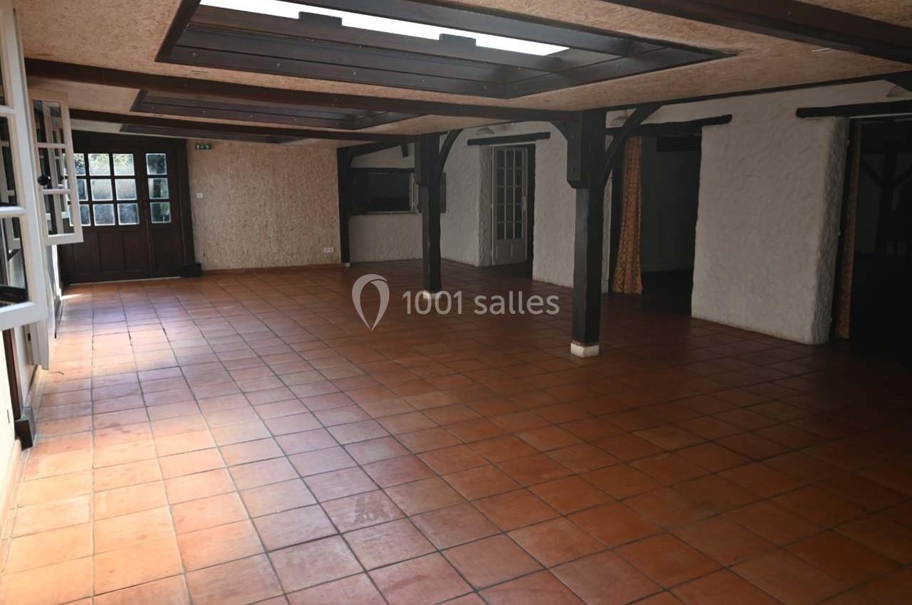Salle spacieuse avec sol en carrelage orange, poutres en bois et grandes fenêtres laissant entrer la lumière naturelle.