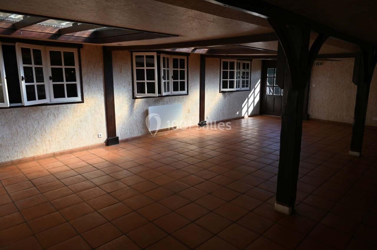 Salle vide avec carrelage en terre cuite, murs clairs, fenêtres à carreaux et poutres en bois apparentes.