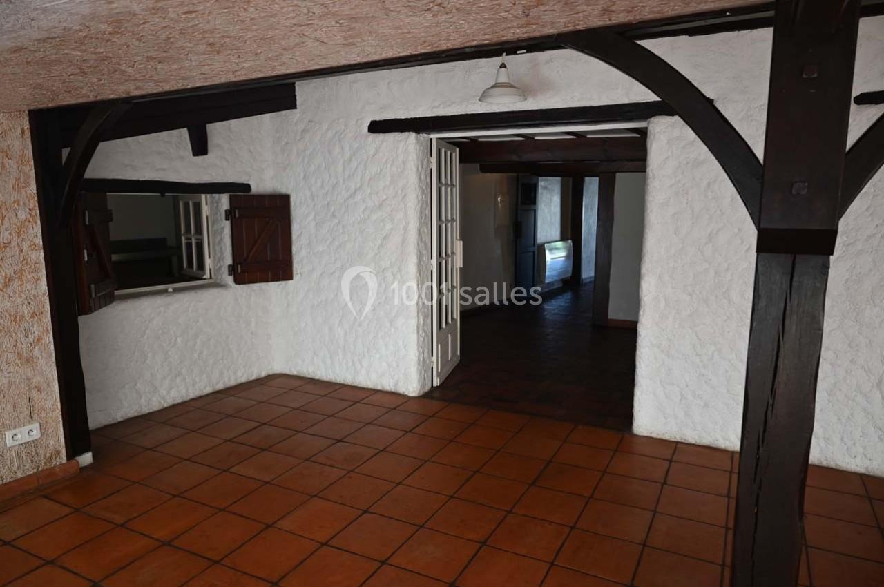 Salle vide avec murs blancs texturés, poutres en bois sombre, carrelage au sol et une ouverture vers une autre pièce.