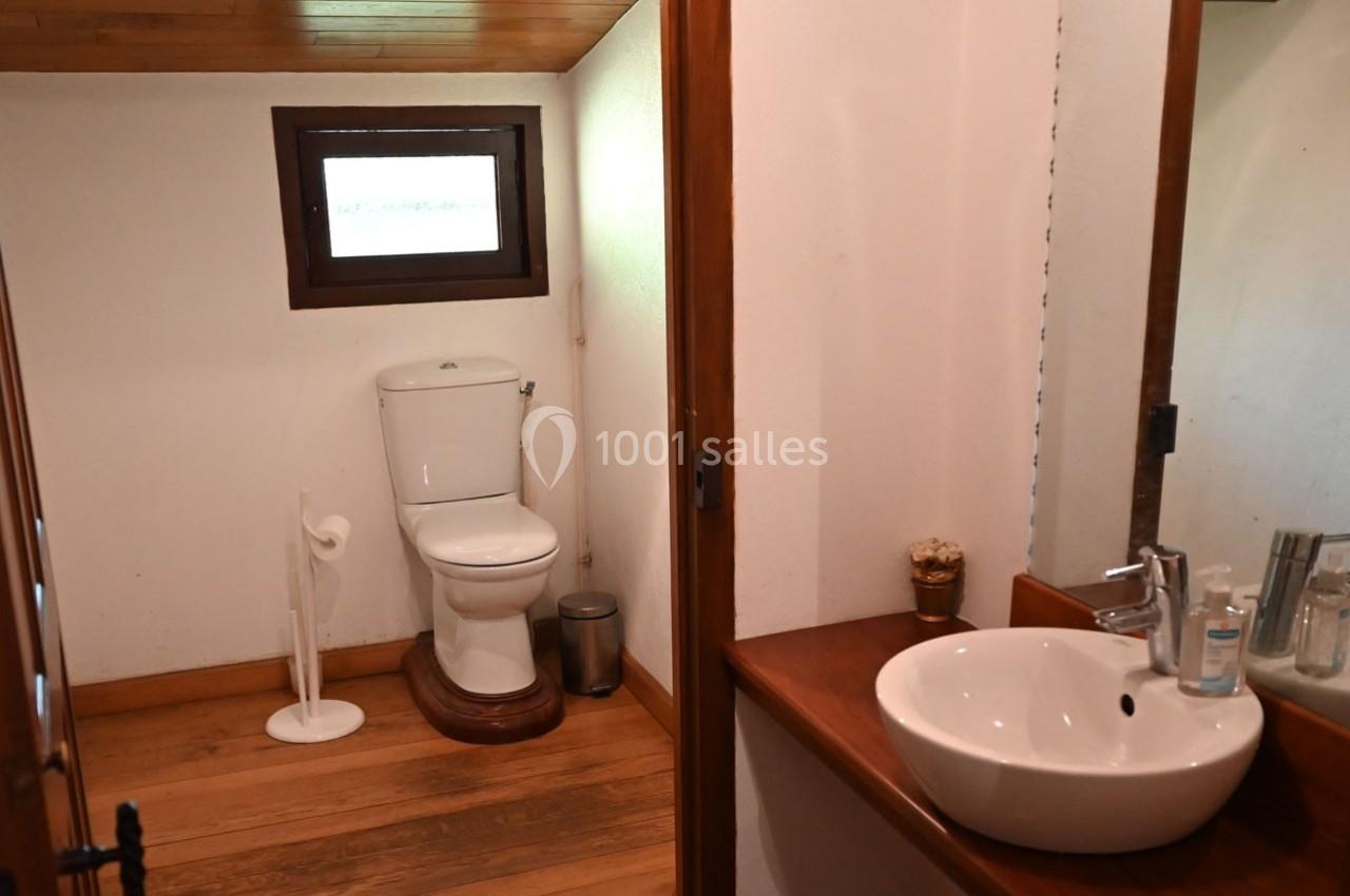 Salle de bain avec toilettes, lavabo blanc, miroir et sol en bois, éclairée par une petite fenêtre rectangulaire.