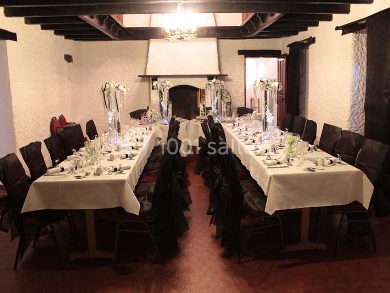 Salle de réception avec deux longues tables dressées, nappes blanches, chaises noires et décorations florales.