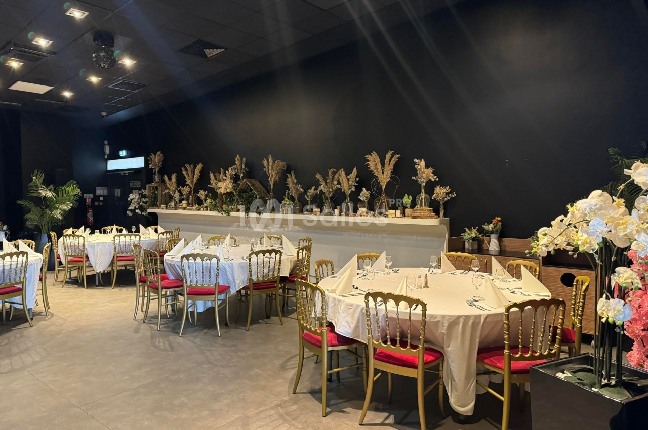 Salle de réception élégante avec tables rondes dressées, chaises dorées et décorations florales sur fond sombre.