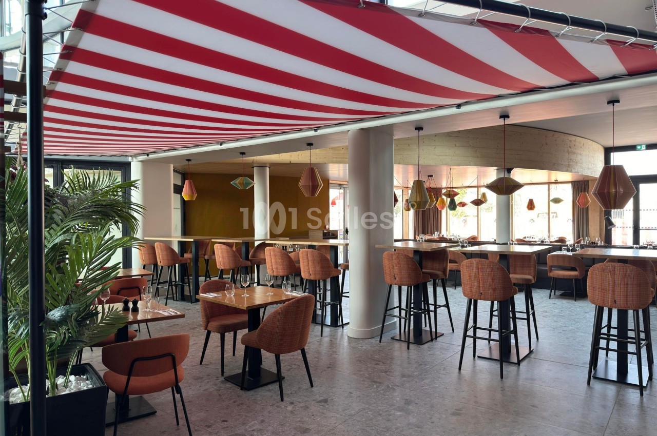 Salle de restaurant moderne avec tables en bois, chaises orange, luminaires suspendus et auvent rayé rouge et blanc.