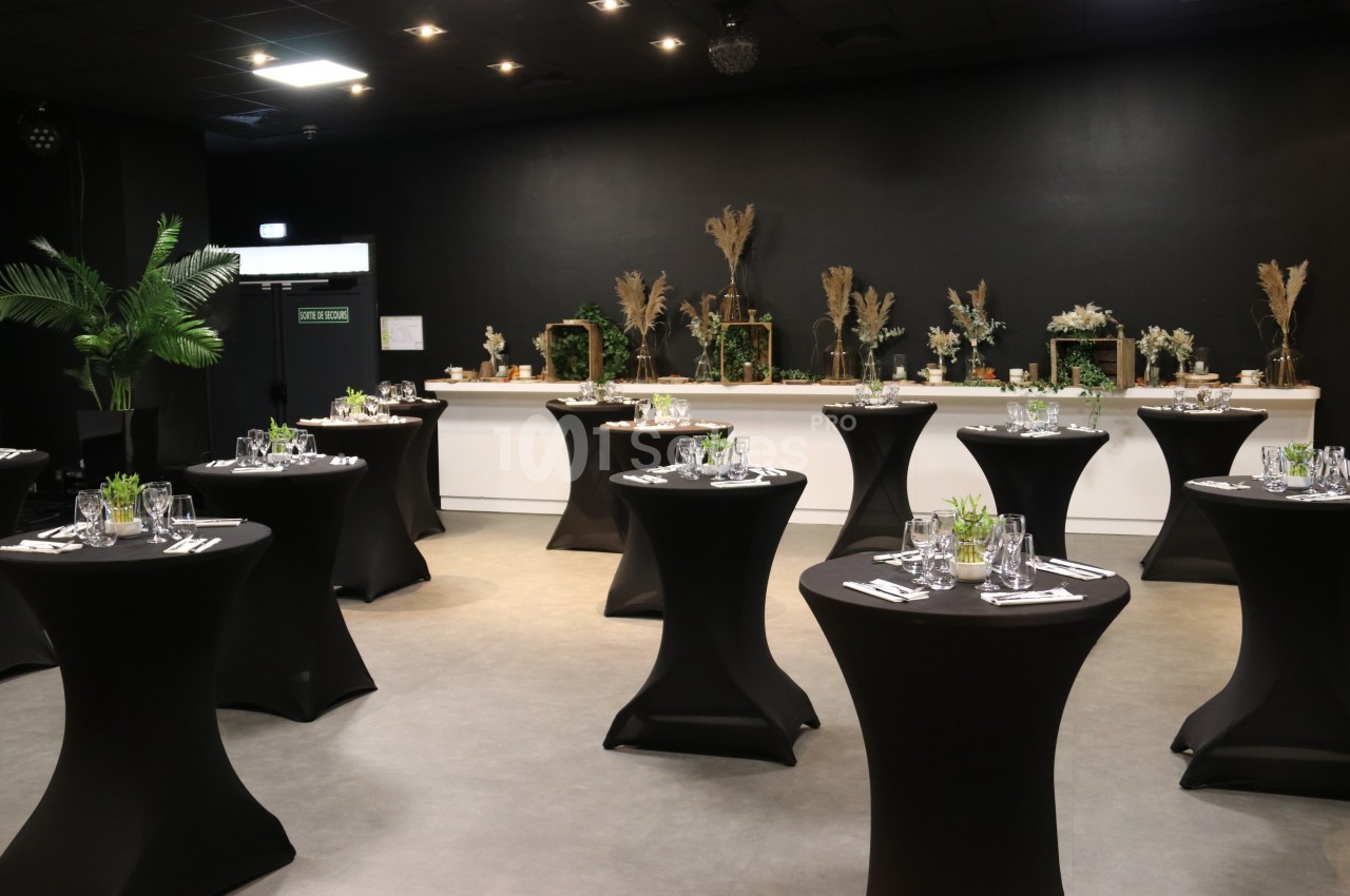 Salle événementielle avec des tables hautes noires dressées, décorations végétales et éclairage tamisé.