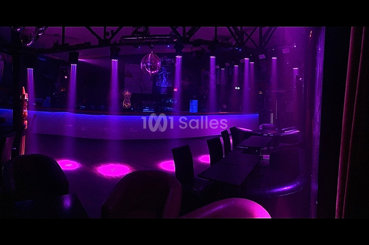 Salle de discothèque vide avec éclairage violet, bar illuminé en arrière-plan et chaises disposées autour de tables.