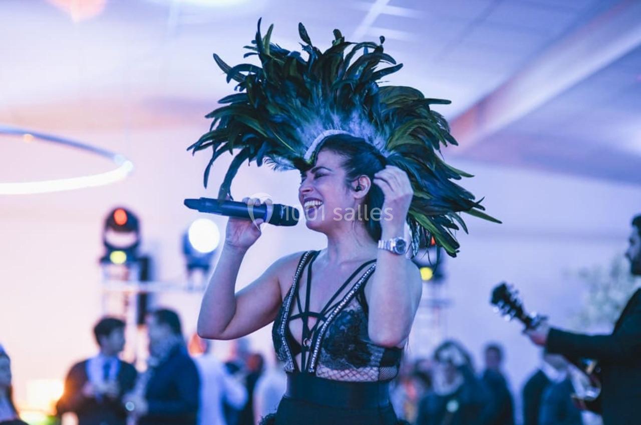 Chanteuse en tenue de scène avec coiffe à plumes, micro à la main, performe devant un public dans une salle éclairée.