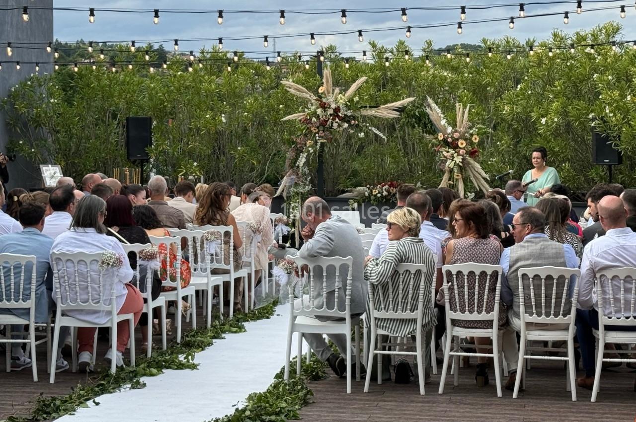 Cérémonie en plein air avec invités assis, allée décorée de feuillage, arche florale et guirlandes lumineuses.