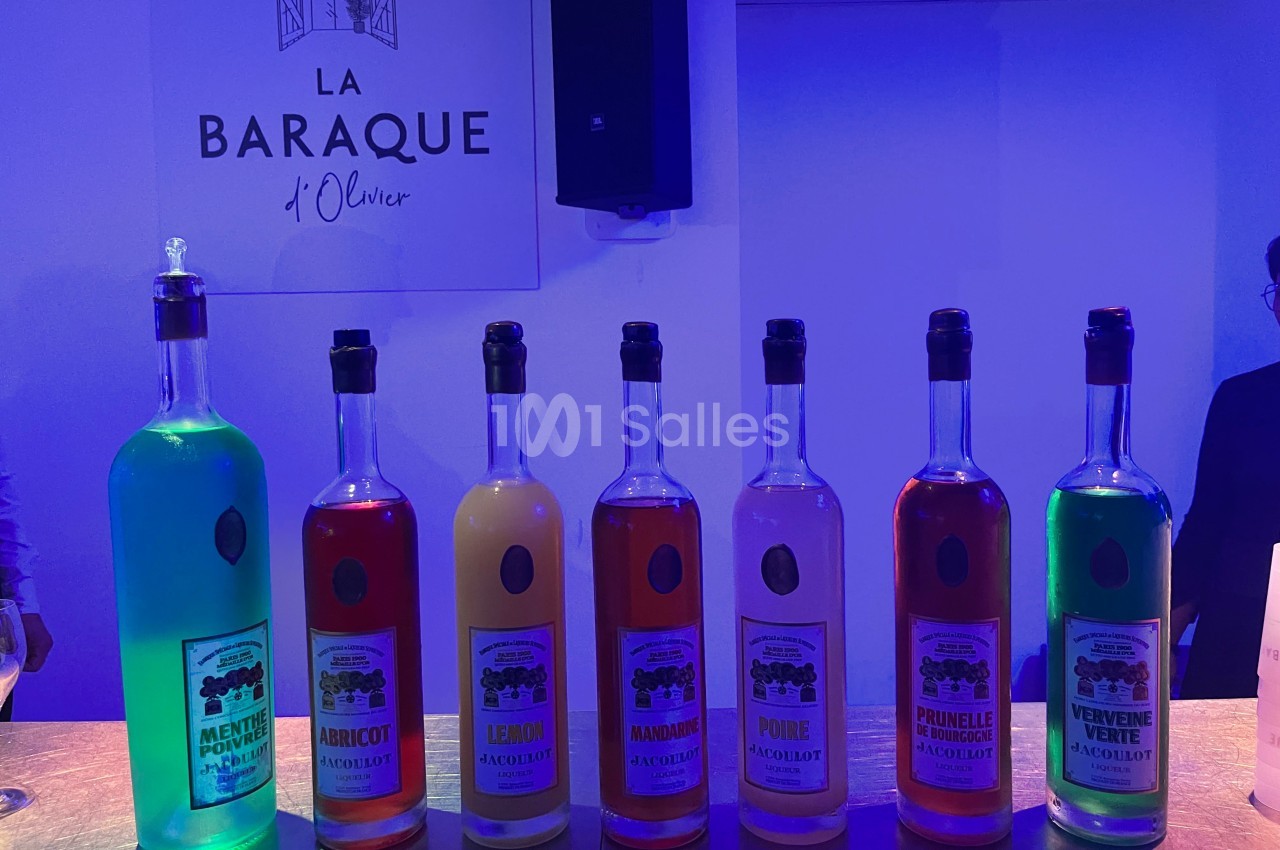 Bouteilles colorées d'apéritifs alignées sur un comptoir, éclairées par une lumière bleutée, avec un panneau en arrière-plan.