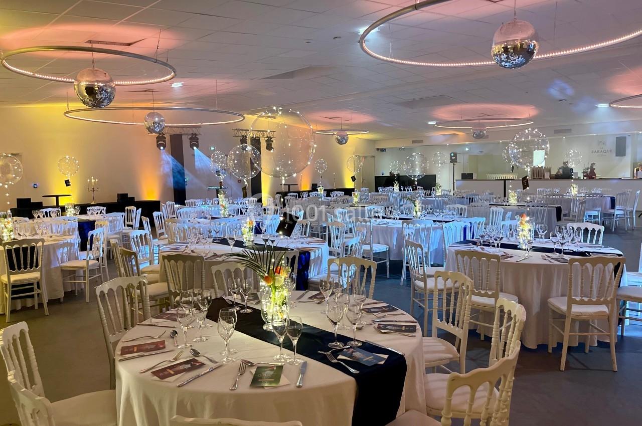Salle de réception décorée avec des tables rondes dressées, éclairage chaleureux et boules lumineuses suspendues.