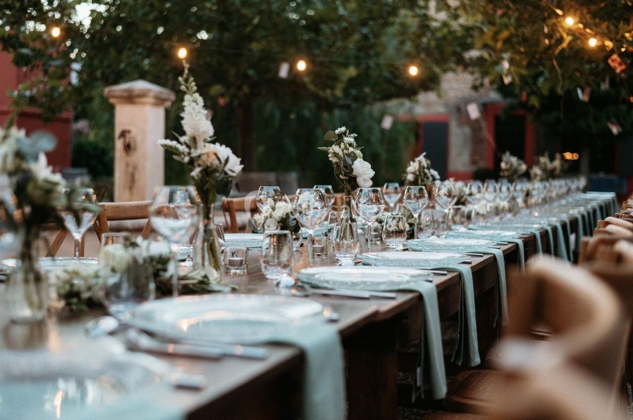 Table dressée en extérieur avec nappes bleues, verres, couverts et décorations florales sous des guirlandes lumineuses.