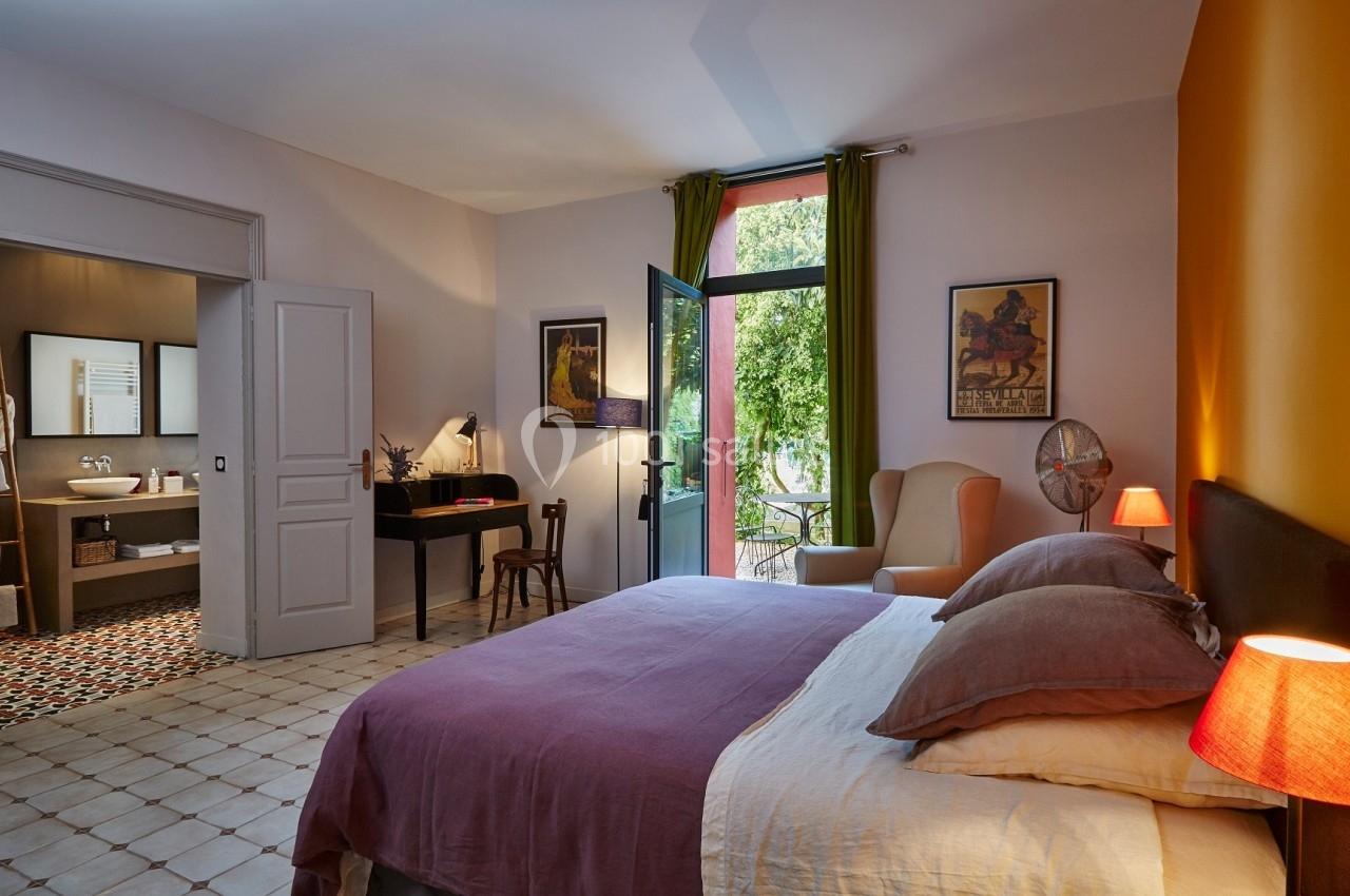 Chambre lumineuse avec lit double, bureau, fauteuil, salle de bain attenante et vue sur un jardin par une porte-fenêtre.