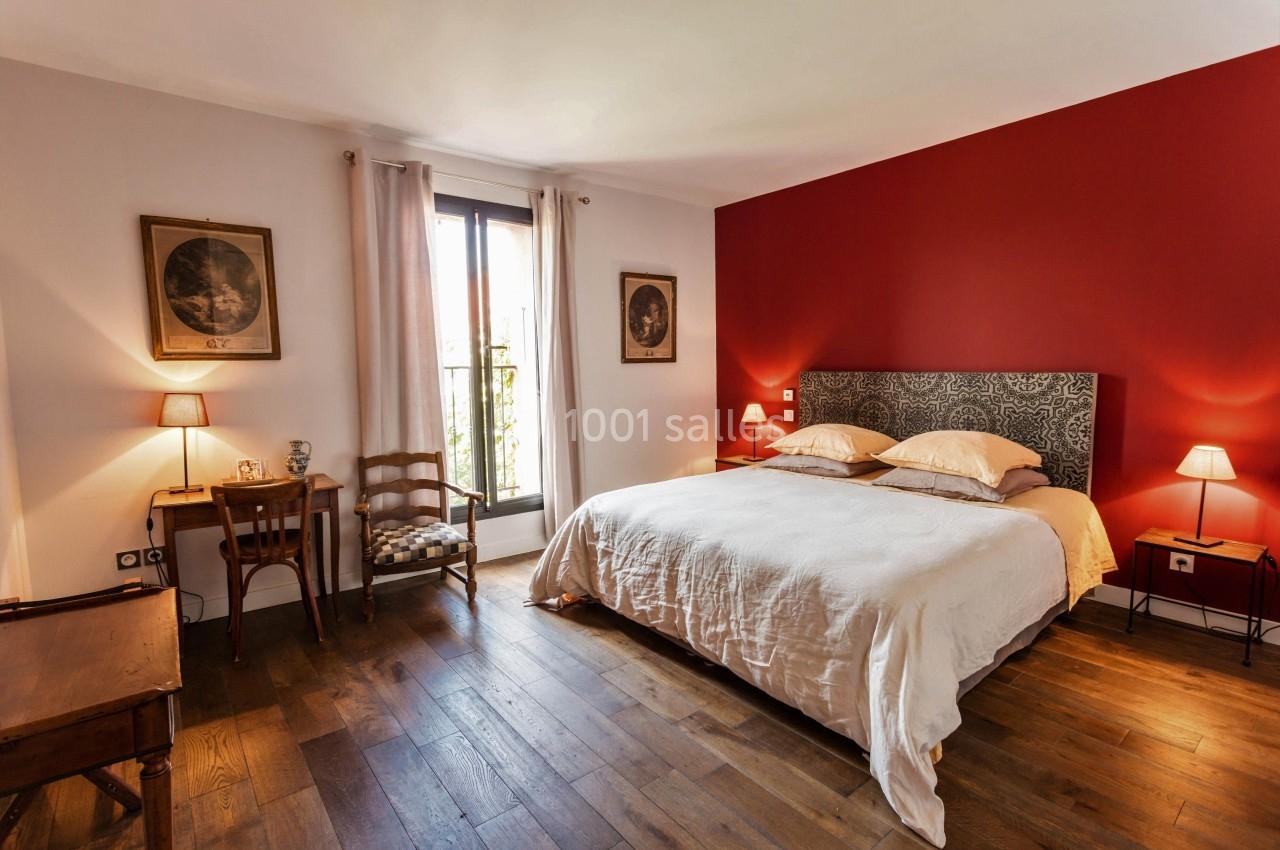 Chambre avec un grand lit, mur rouge, parquet en bois, bureau, chaise et fenêtre laissant entrer la lumière naturelle.