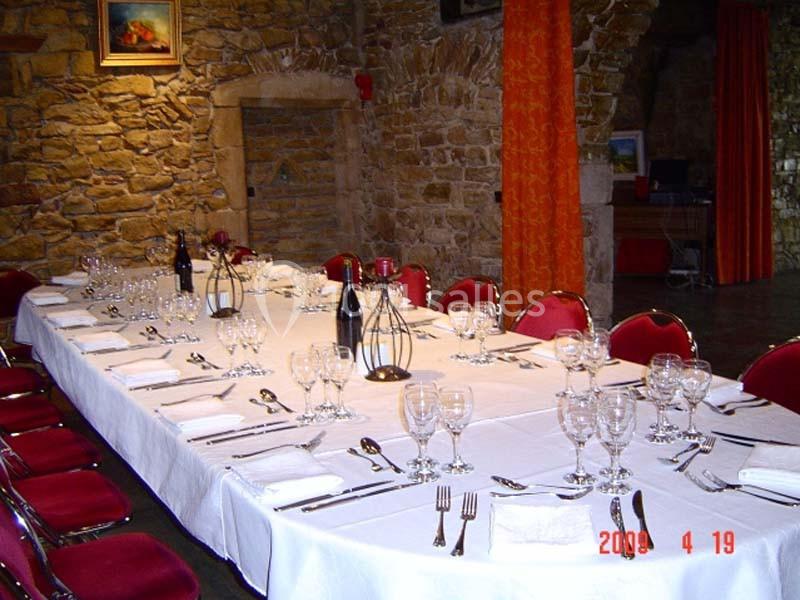 Table longue dressée avec nappes blanches, verres, couverts et chaises rouges, dans une salle aux murs en pierre.