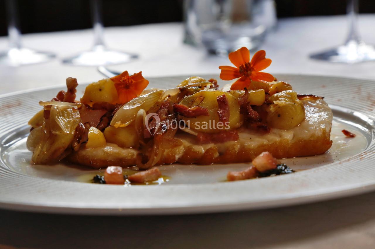 Tranche de pain garnie de pommes de terre, lardons, oignons et décorée de fleurs comestibles sur une assiette blanche.
