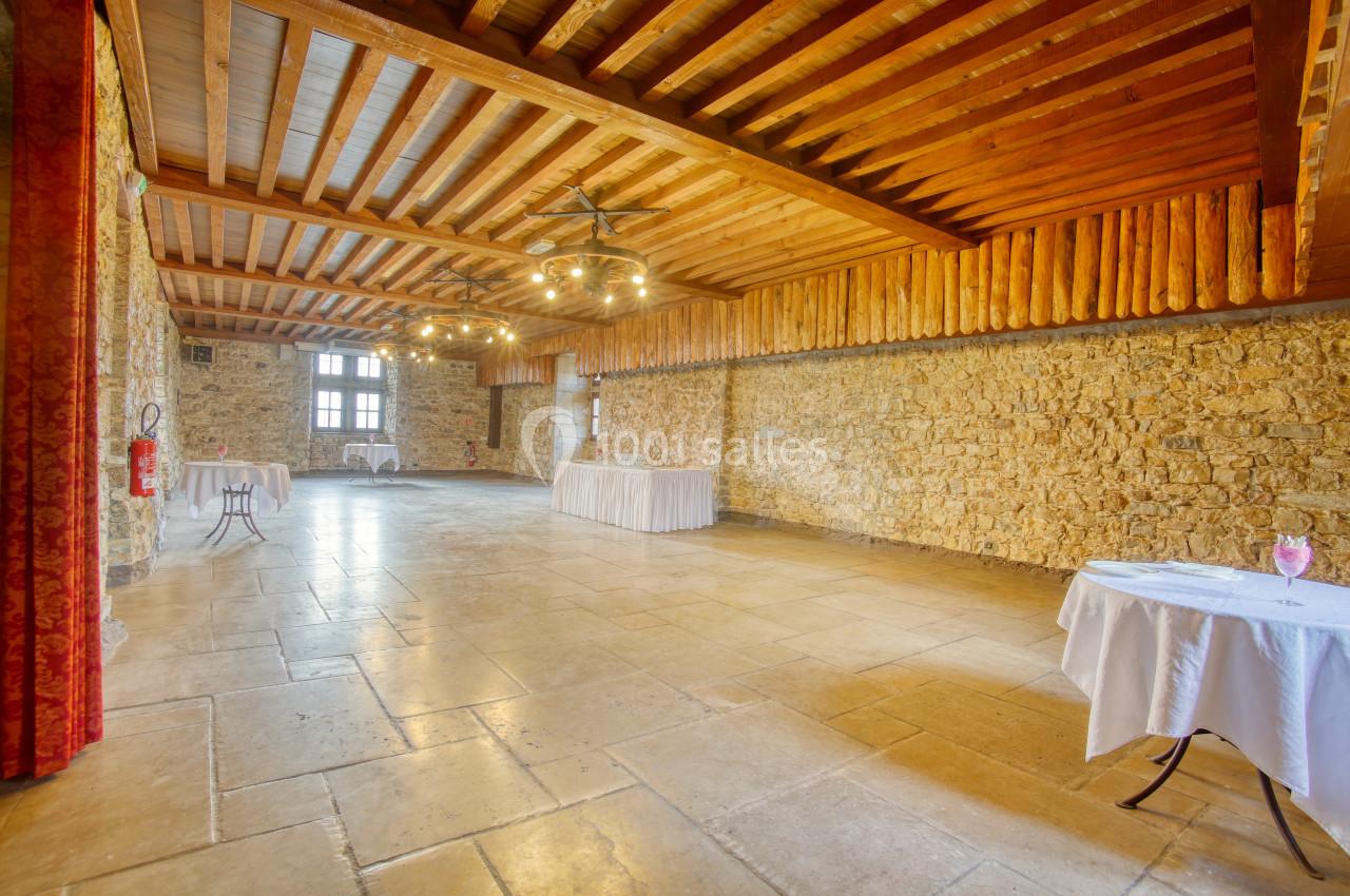 Salle spacieuse avec murs en pierre, plafond en bois et tables rondes dressées, éclairée par des lustres et une grande…