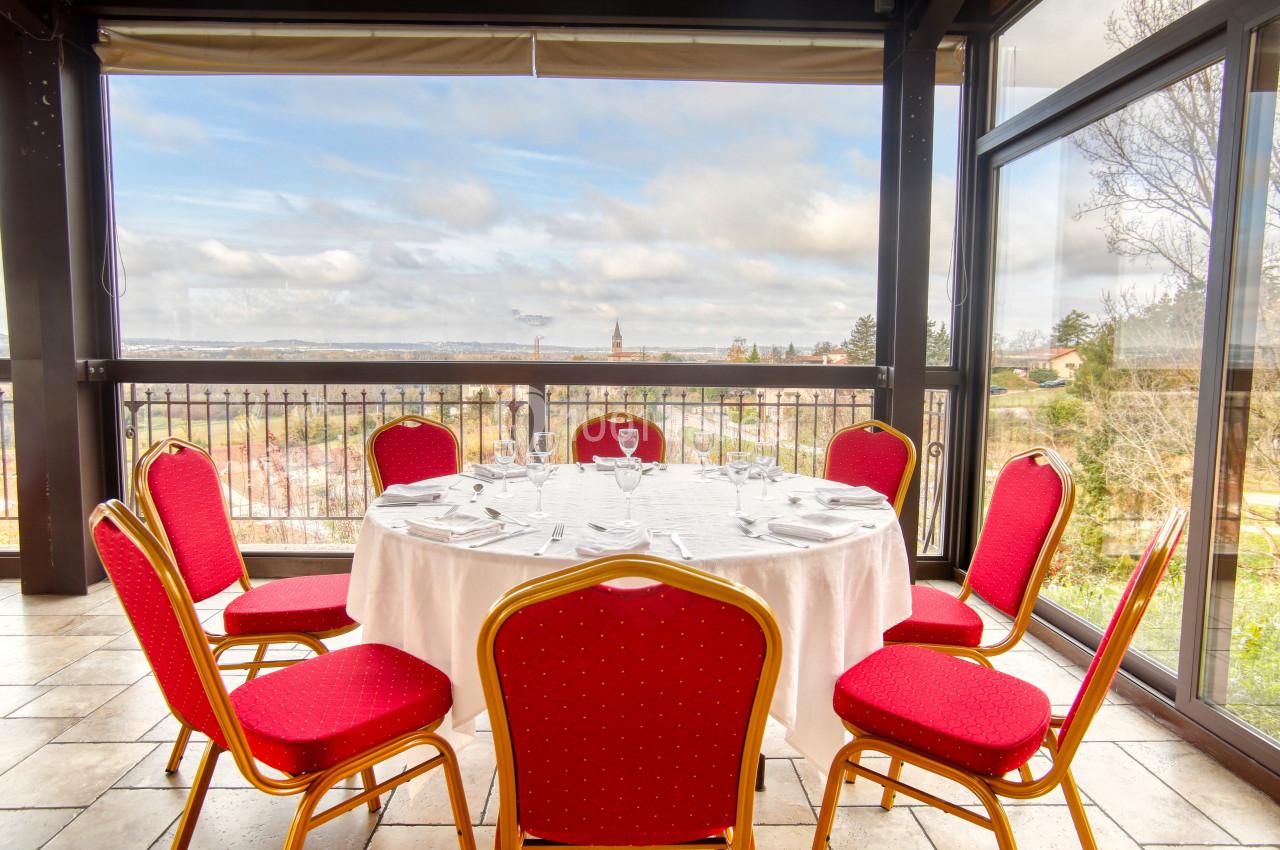 Salle lumineuse avec une table ronde dressée, entourée de chaises rouges, offrant une vue panoramique sur l'extérieur.