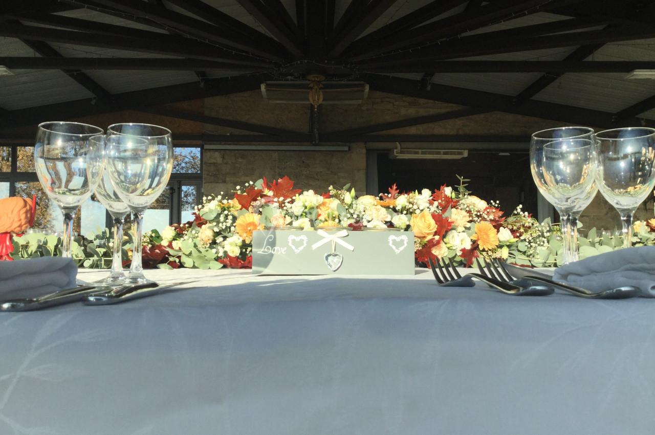 Table décorée avec nappes grises, verres, couverts et centre de table floral aux tons chauds dans une salle lumineuse.