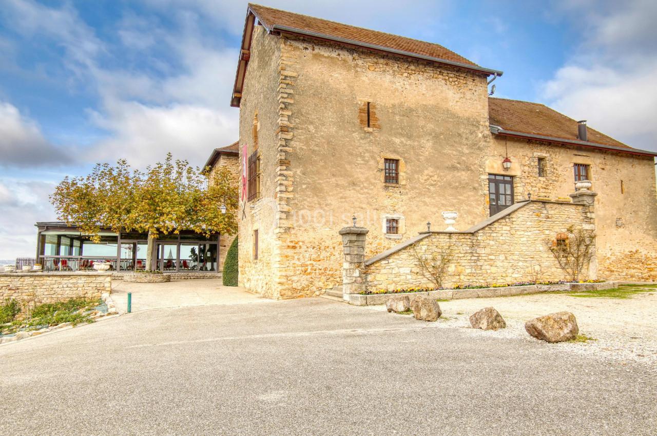 Bâtiment en pierre ancienne avec toit en tuiles, entouré d'arbres et d'une terrasse moderne sous un ciel partiellement…