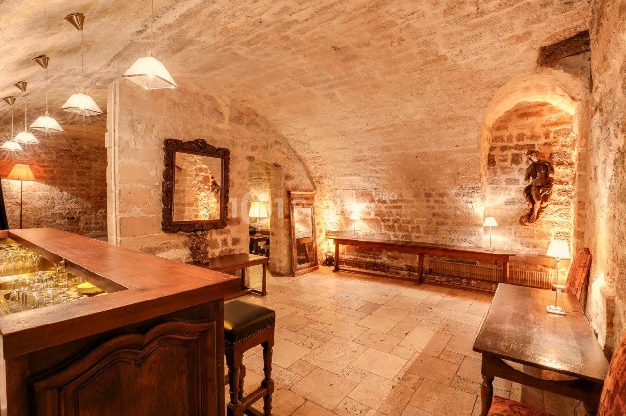 Salle voûtée en pierre avec un bar en bois, des tables, des chaises et un éclairage chaleureux.