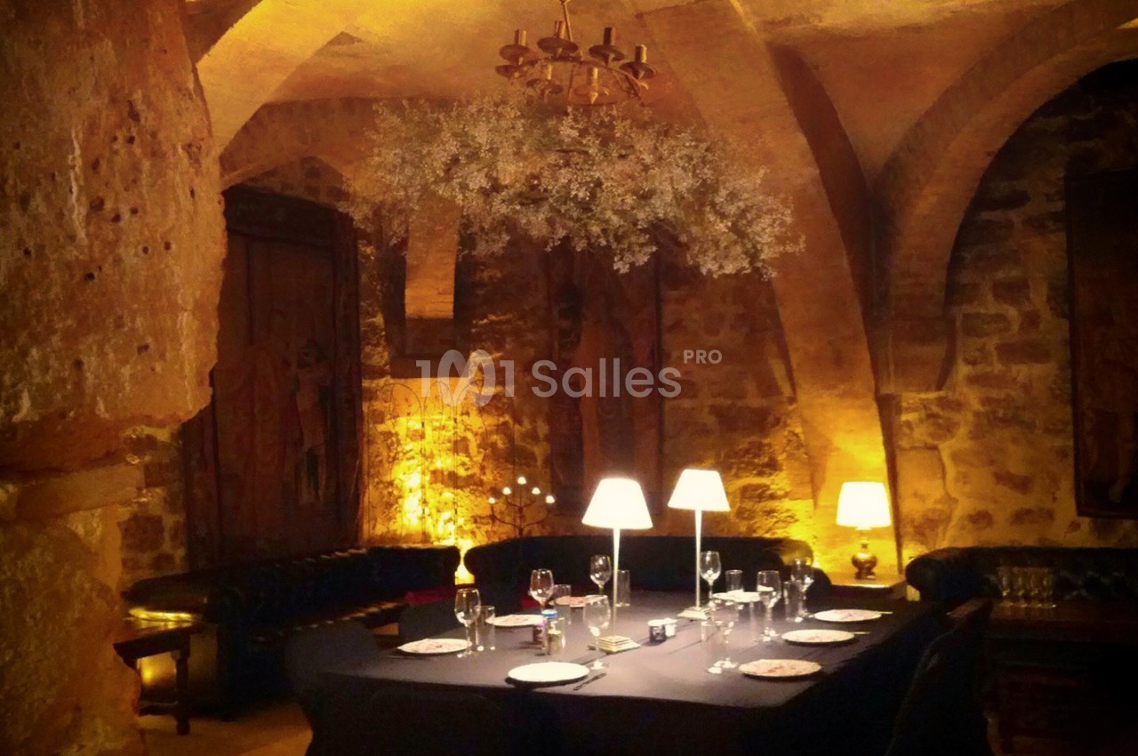 Salle voûtée en pierre éclairée par des lampes et un lustre, avec une table dressée pour un repas.