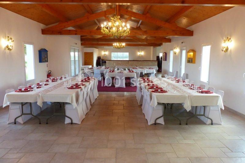 Salle de réception aménagée avec de longues tables dressées, nappes blanches et assiettes rouges, sous un plafond en bois.