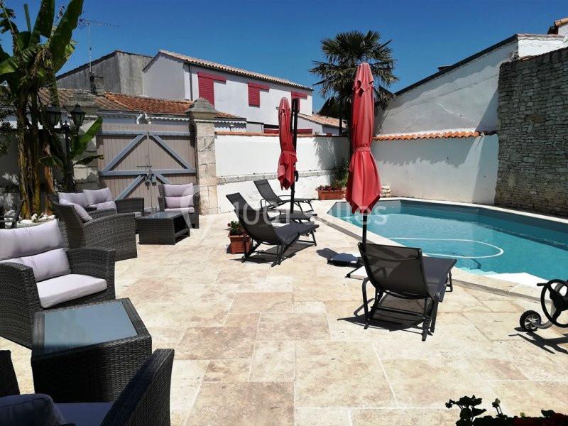 Terrasse ensoleillée avec piscine, transats, parasols rouges, fauteuils en rotin et murs en pierre blanche.