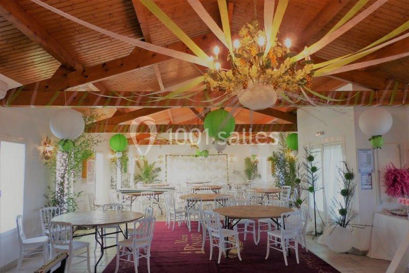 Salle décorée pour un événement avec des tables rondes, des chaises blanches, des guirlandes colorées et des lanternes…