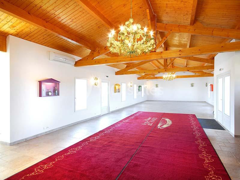 Salle lumineuse avec plafond en bois, lustres dorés et tapis rouge au sol, entourée de murs blancs.