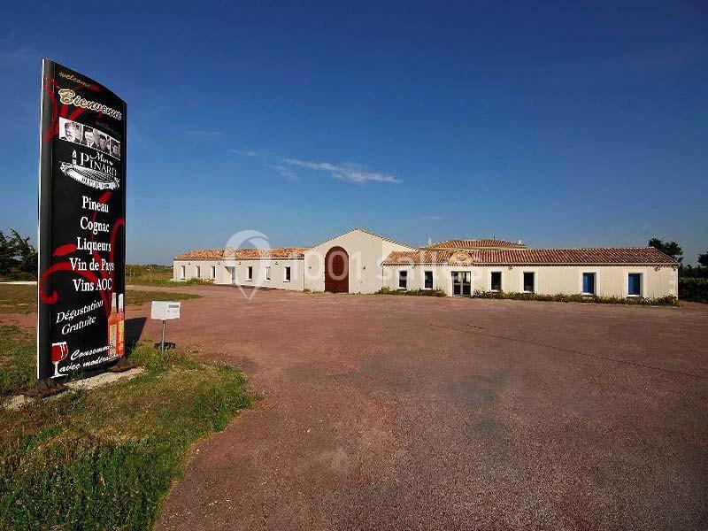 Bâtiment clair avec toit en tuiles entouré d'un grand parking, panneau indiquant une boutique de vins et produits locaux.